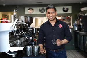 Friendly barista with salt-and-pepper hair smiles while holding a glass espresso cup in front of a coffee machine and metal pitchers.