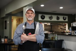 A smiling chef with a white cap and black apron holds a large spoon and a sharp knife in a restaurant setting.